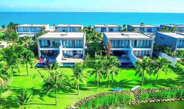 A modern resort with multiple two-story buildings, each featuring balconies and large windows. The resort is surrounded by lush greenery and palm trees, with a view of the ocean. - Sanctuary Ho Tram Resort – Private Pool Villas (Ho Chi Minh City)