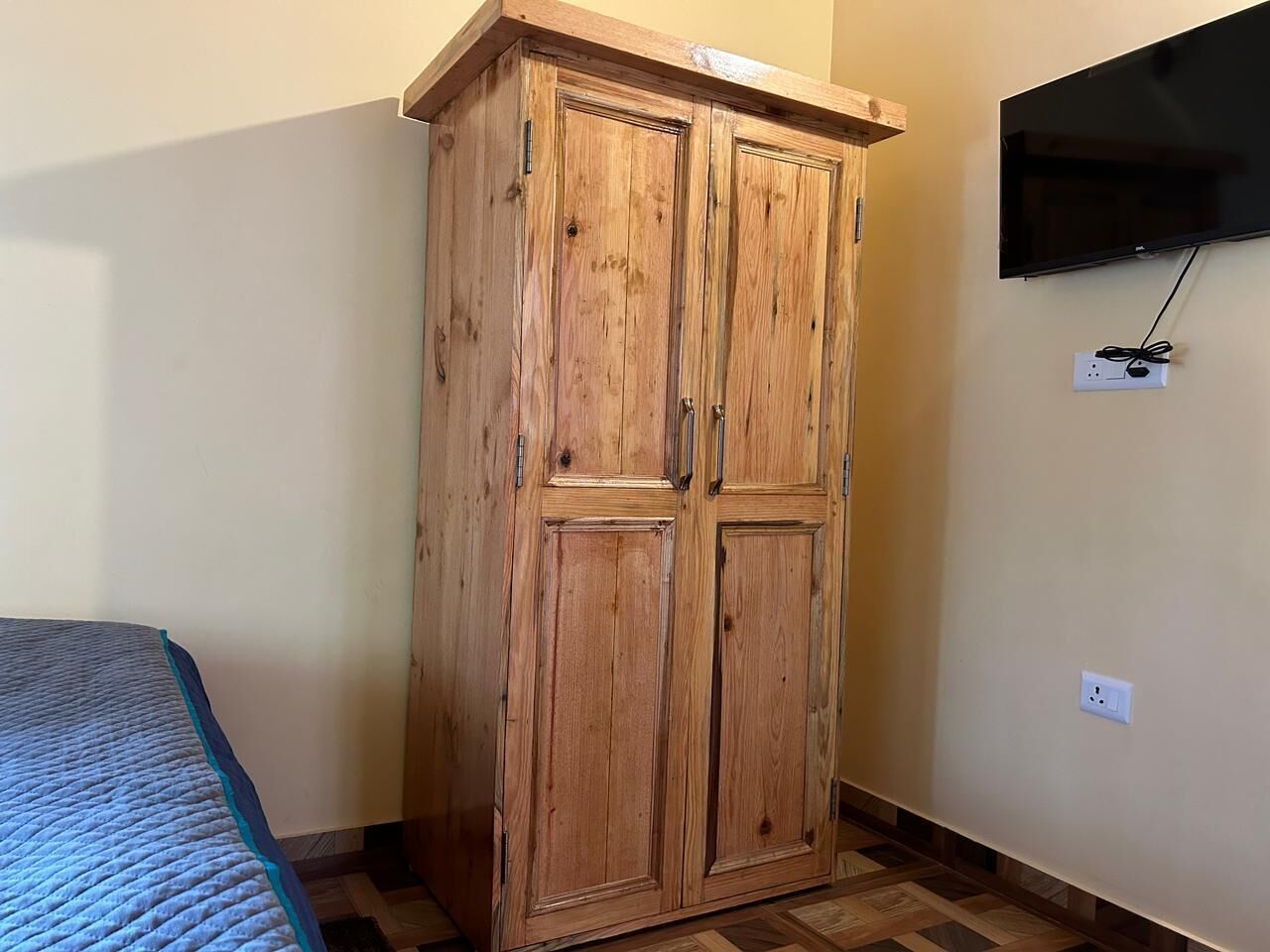 A room with a wooden wardrobe, a bed with a blue quilt, and a mounted TV on the wall.