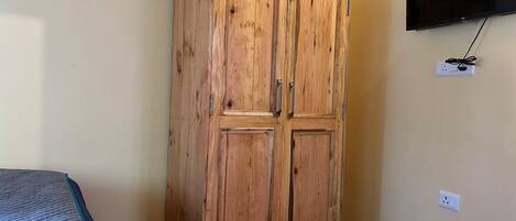A room with a wooden wardrobe, a bed with a blue quilt, and a mounted TV on the wall.