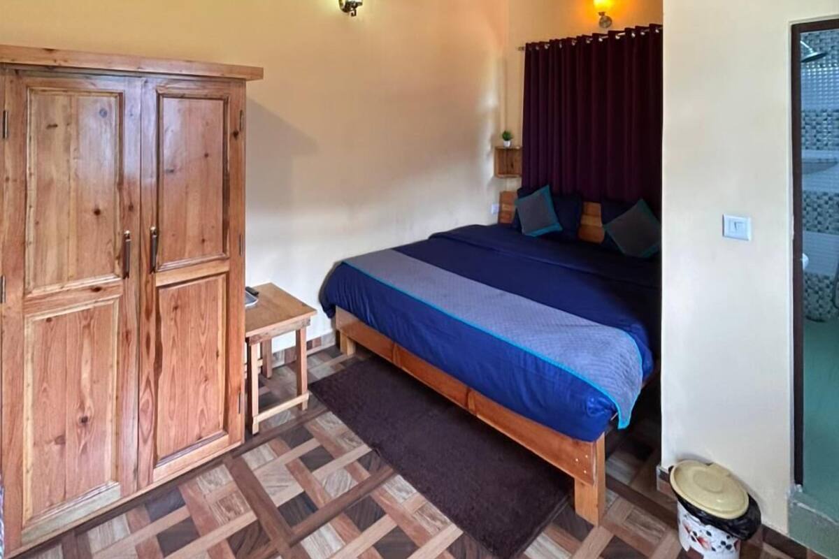 Un dormitorio con techo de madera, una cama con colcha azul, un armario de madera, una mesita y un suelo estampado.