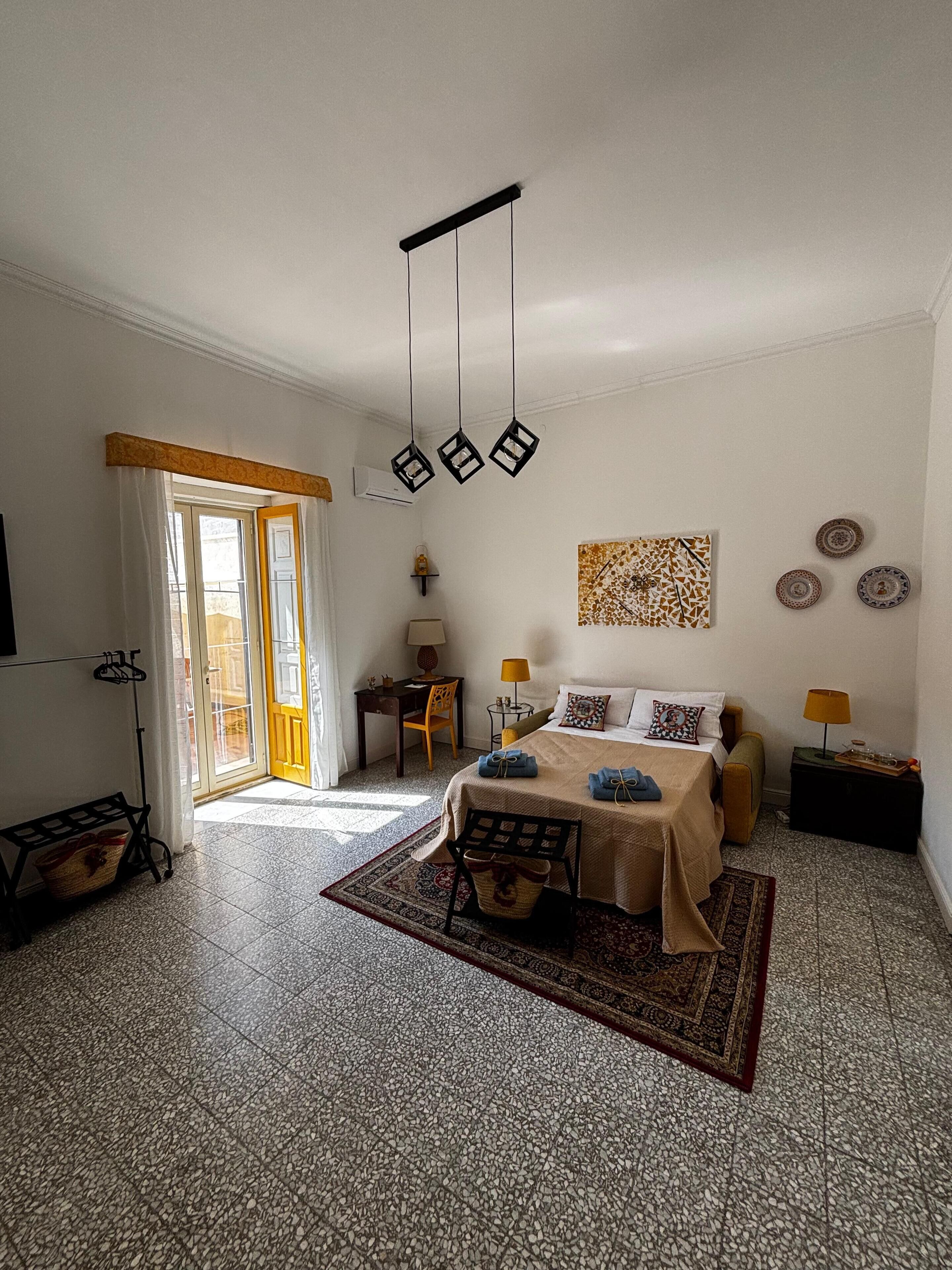Une chambre d’hôtel comprenant un lit, un bureau, une chaise, une petite table avec une lampe, une étagère fixée au mur et une fenêtre avec des rideaux.