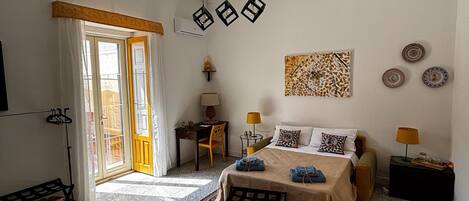 Une chambre d’hôtel comprenant un lit, un bureau, une chaise, une petite table avec une lampe, une étagère fixée au mur et une fenêtre avec des rideaux.