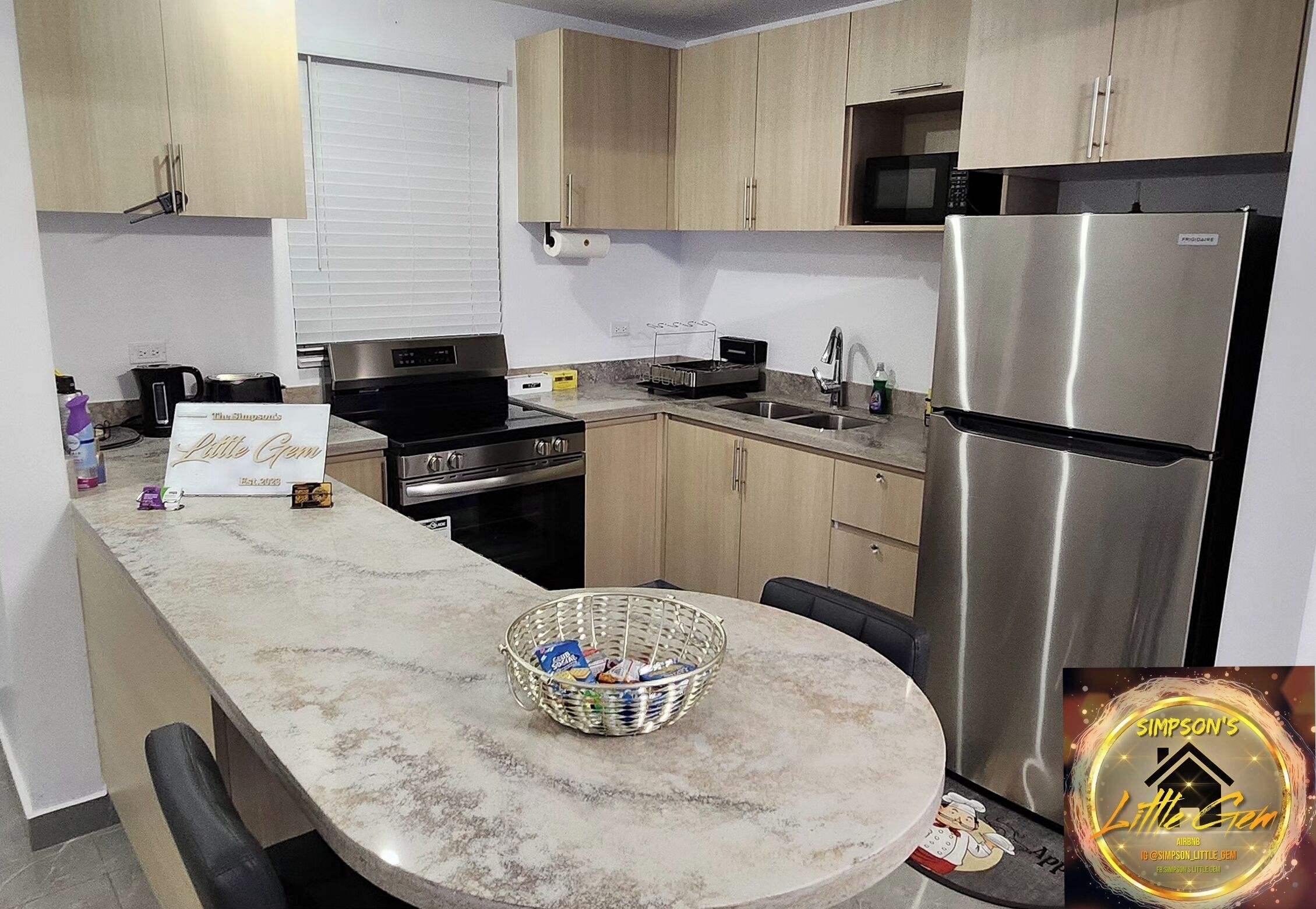 Une cuisine moderne avec des meubles en bois clair, un réfrigérateur en acier inoxydable, un four à micro-ondes et une cuisinière à gaz. On y trouve un plan de travail en marbre, un panier rempli d’en-cas et une pancarte indiquant « Little Gem ».