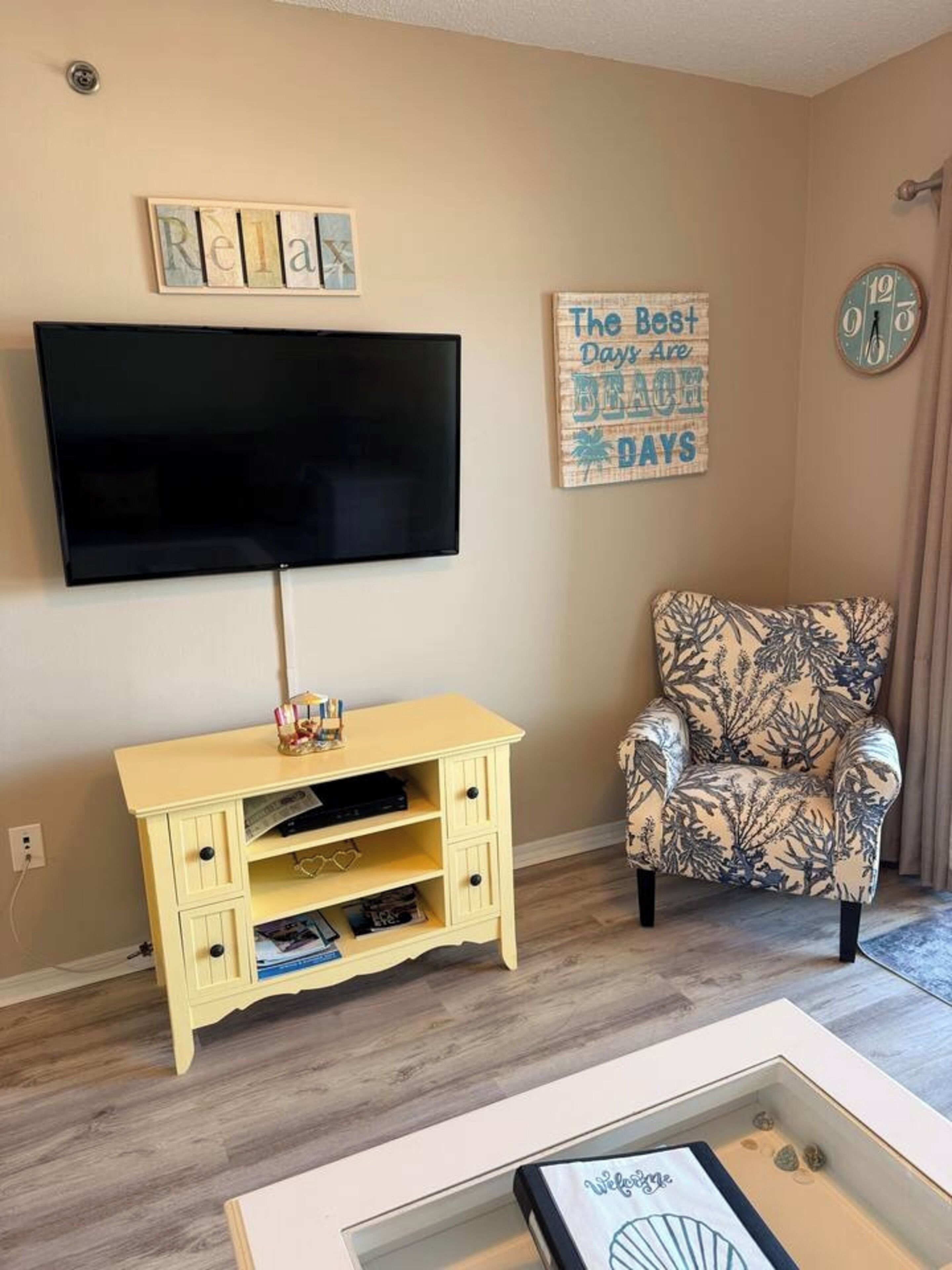 A living room with a flat-screen TV, a yellow storage unit, a patterned armchair, and a wall clock.