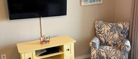 A living room with a flat-screen TV, a yellow storage unit, a patterned armchair, and a wall clock.