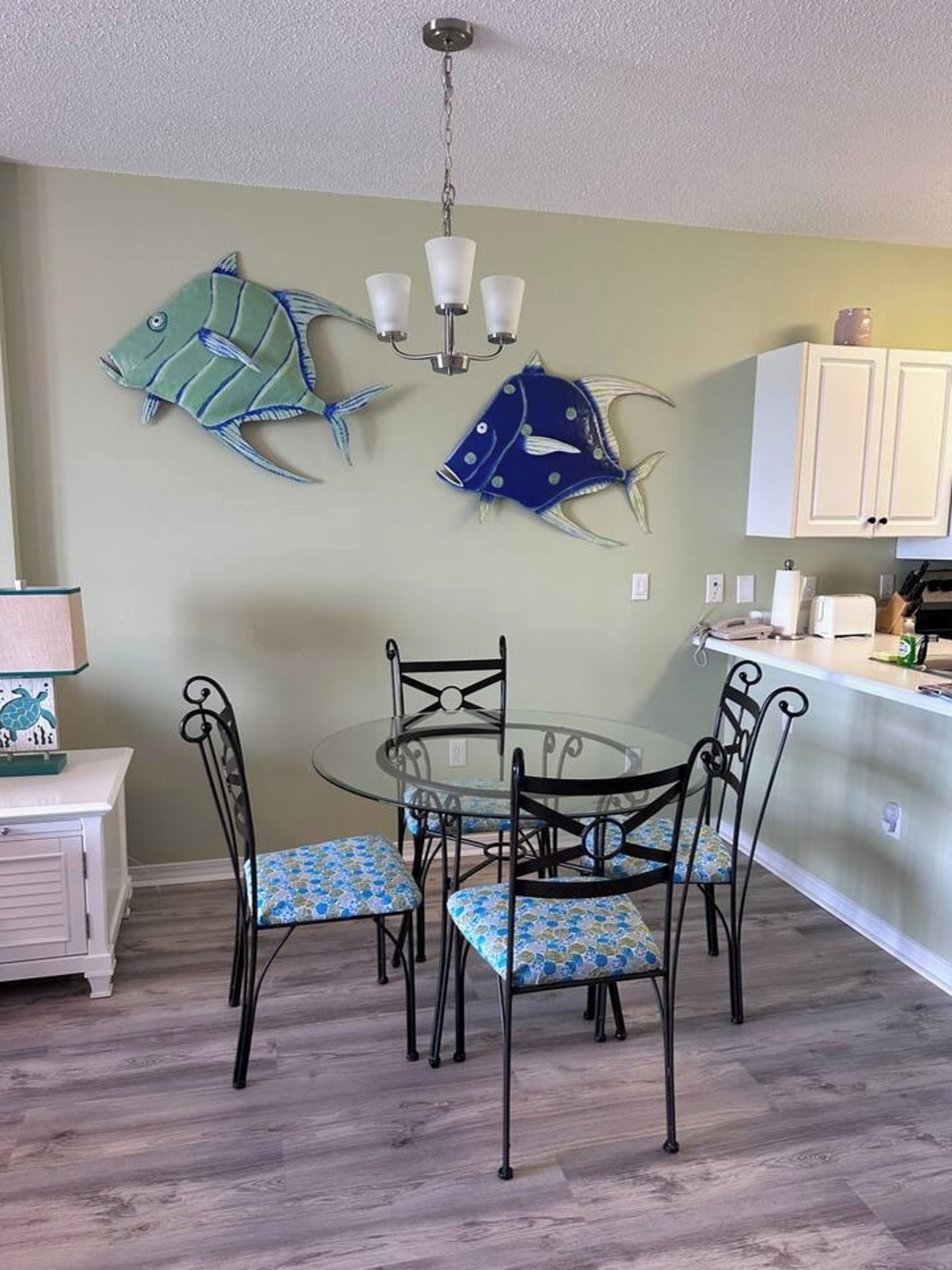 Un comedor con una mesa de vidrio y sillas de metal, una araña y un arte mural decorativo con motivos de peces.