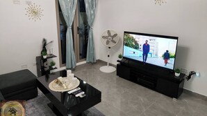 A modern living room with a flat-screen TV, a black entertainment unit, a glass door with curtains, a standing fan, and a coffee table with decorative items. - Simpson's Little Gem 1 (Kingston)