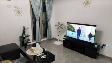 Una sala de estar moderna con televisor de pantalla plana, mueble de entretenimiento negro, puerta de cristal con cortinas, un ventilador de pie y una mesa de centro con adornos.