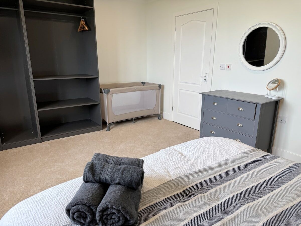 A bedroom with a bed, a grey dresser, a wardrobe, a bench, and a round mirror.