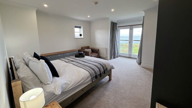 A modern bedroom with a large bed, bedside tables, a window with curtains, and a view of the outdoors. - Peg-y-Don : Stunning Holiday Cottage in Newport, Pembrokeshire (Newport)