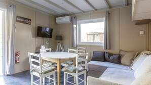 A room with a round table, white chairs, a sofa, a TV, and a window with curtains. - Chalet 809 with all comforts on the edge of the forest (La Teste-de-Buch)