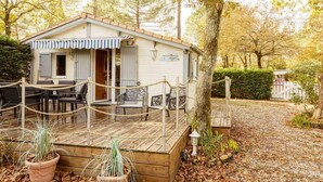 A small, single-story cabin with a deck, surrounded by trees and a gravel path. - Chalet 213 on the edge of a forest (La Teste De Buch)