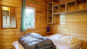 A wooden cabin room with a bed, a window with curtains, and built-in shelves. - Chalet 213 on the edge of a forest (La Teste De Buch)