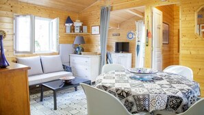 A cozy cabin with a wooden interior, a dining area with a patterned tablecloth, a sofa, and a small kitchen area. - Chalet 213 on the edge of a forest (La Teste De Buch)
