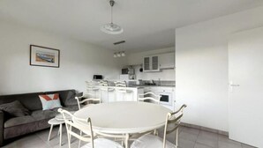 A modern living room with a grey sofa, a round white table, and white chairs. A kitchen with white cabinets and a white refrigerator is visible in the background. - (P) T2 2 steps from the Bassin (Gujan-Mestras)