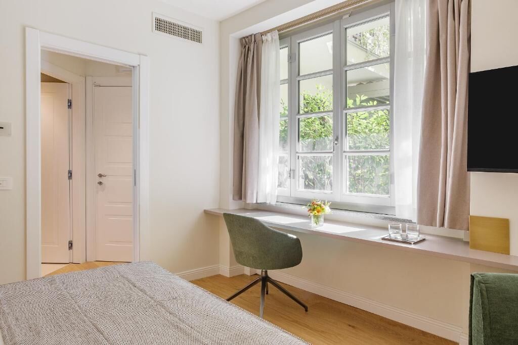 A bedroom with a bed, a desk with a chair, a window with curtains, and a TV on the wall.
