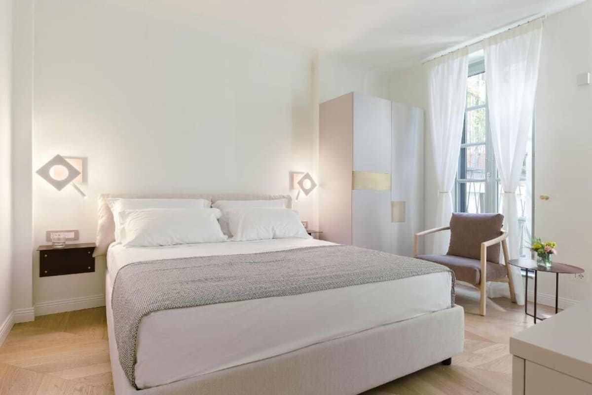 A modern bedroom with a large bed, a chair, a small table, and a window with curtains.
