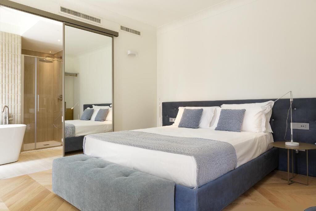 A modern bedroom with a large bed, a bedside table, a mirror, and a bathroom with a bathtub.