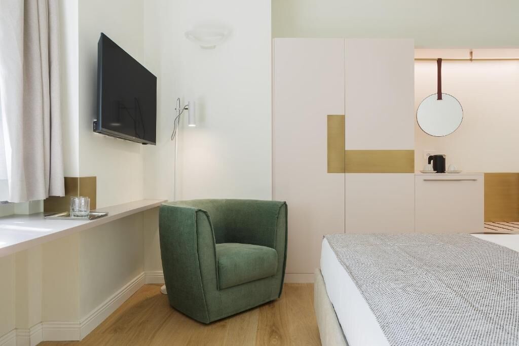 Una camera da letto moderna con una poltrona verde, un letto con copripiumino fantasia, una TV a parete, uno specchio e un tavolino con un bollitore.