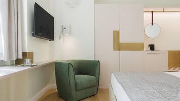 Una camera da letto moderna con una poltrona verde, un letto con copripiumino fantasia, una TV a parete, uno specchio e un tavolino con un bollitore.