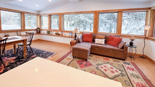 A cozy living room with a wooden dining table, a sofa with red cushions, and large windows offering a view of a snowy forest.