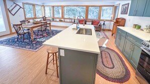 A modern kitchen with a central island, wooden dining table, and large windows offering a view of snow-covered trees.