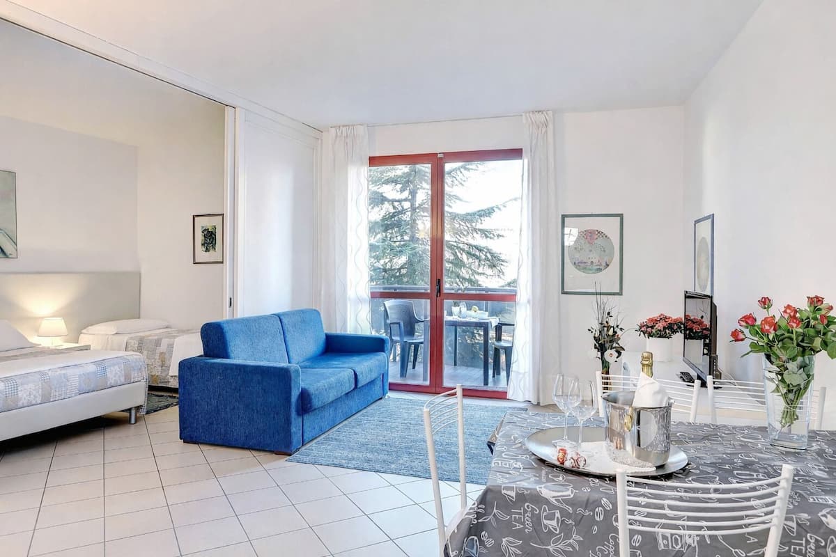 Habitación de hotel moderna con un sofá azul, una mesa de comedor con mantel de pizarra y un balcón con vistas a árboles.