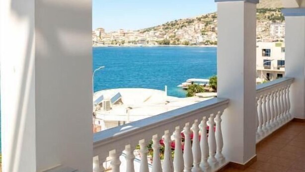 A balcony with a white balustrade overlooking a coastal area with buildings and a clear blue sea. - Erdano Hotel (Sarande)