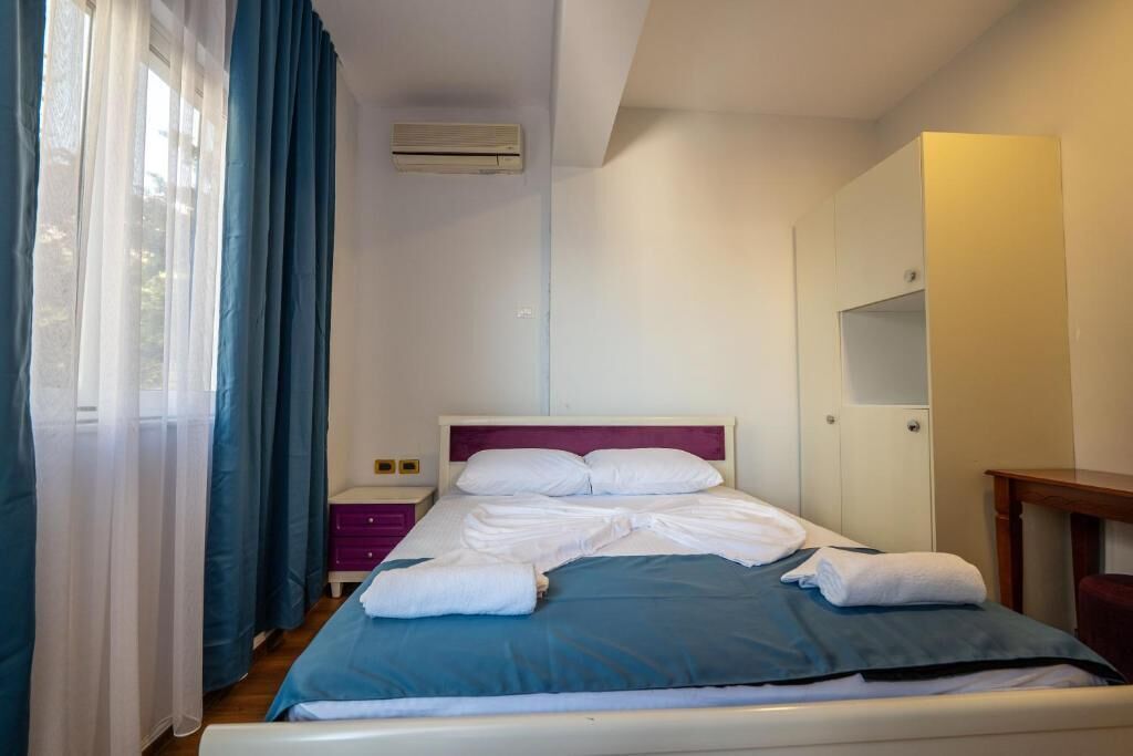 Una cama bien hecha con sábanas blancas y una manta azul, una mesita de noche, una mesita de cabecera morada y una ventana con cortinas azules.
