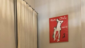 En snyggt bäddad säng med vita lakan, en hopvikt handduk och en liten tygpåse med dragsko på vänster sida. En affisch med en kvinna och en hund hänger på väggen ovanför sängen.