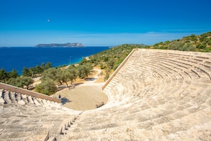 Ein Amphitheater mit terrassenförmigen Sitzreihen, freiem Blick auf das Meer und einer hügeligen Landschaft im Hintergrund.