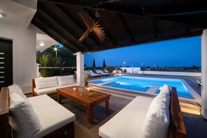 Ein Poolbereich mit Holztisch, weiß gepolsterten Sitzgelegenheiten und Blick auf den Pool im Abenddämmerung.
