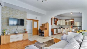 Una sala de estar moderna con sofá, una mesa de centro y televisor. Se observa una zona de cocina con armarios y una mesa de comedor.