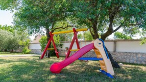 Un parque infantil con un sube y baja rojo y amarillo, y un tobogán, rodeado de césped y árboles.