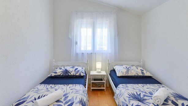Una habitación pequeña y limpia con dos camas individuales, una mesita de noche con lámpara y una ventana con cortinas traslúcidas.