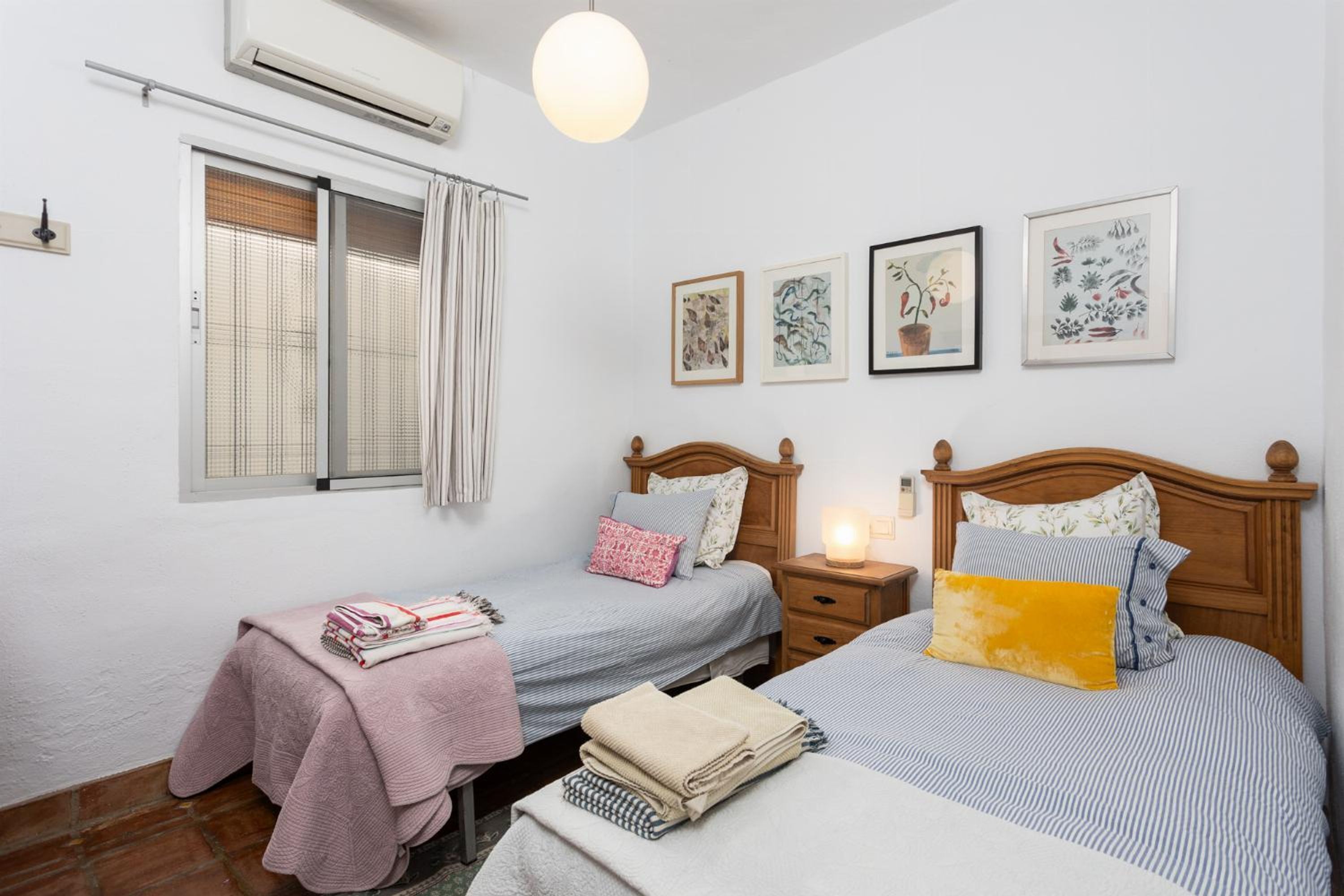 A bedroom with two beds, a window with curtains, a bedside table with a lamp, and wall art.