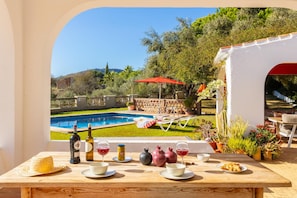 Ein Esstisch mit Weinflaschen, Gläsern und Granatäpfeln, mit Blick auf einen Swimmingpool und eine Außenbereich-Sitzgruppe mit einem roten Sonnenschirm.