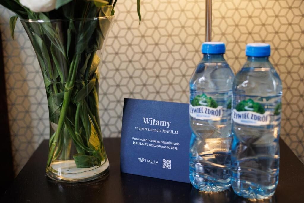 Eine Glasvase mit grünen Stielen und Blättern, zwei Wasserflaschen mit blauen Verschlusskappen und ein Schild mit Text in einer Fremdsprache.