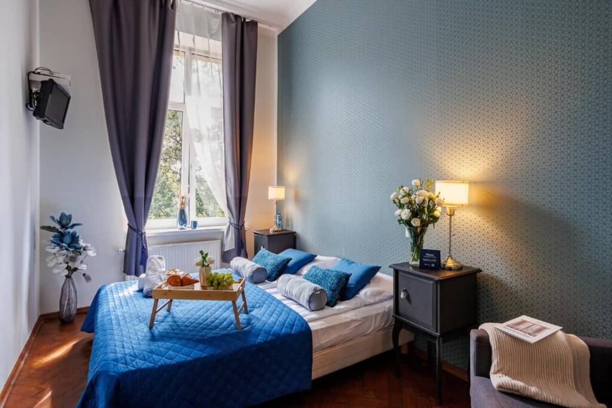 Ein Schlafzimmer mit einem Bett, Nachttischen, Vorhängen am Fenster, einer Vase mit Blumen sowie einem Tablett mit Früchten und einem Buch.