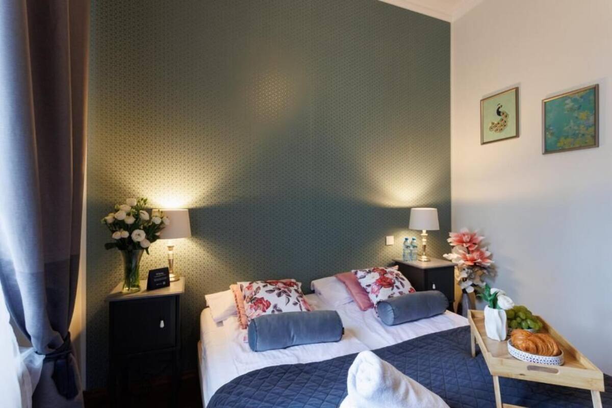 Ein Hotelzimmer mit einem Bett, zwei Nachttischen, einer Ablagefläche, einer Schale mit Früchten und Gebäck sowie einer Blumenvase.