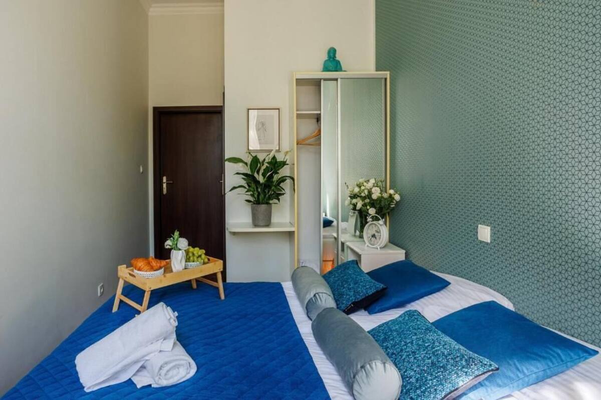 Ein Schlafzimmer mit Bett, Nachttisch, Spiegel, Kleiderschrank, einer kleinen Pflanze und einer Ablage mit Früchten und einem Handtuch.