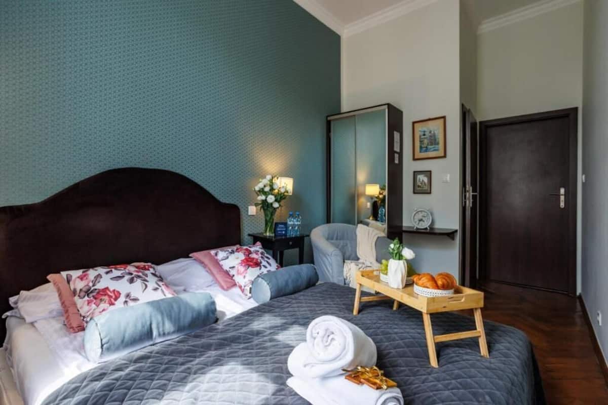 Ein ordentlich bezogenes Bett mit gesteppter Decke, ein Nachttisch mit einer Platte mit Brot und einer Flasche, ein Spiegel und eine Tür in einem Hotelzimmer.