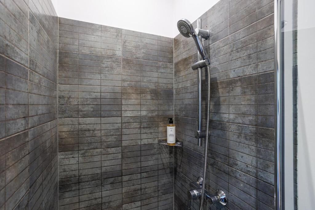 Eine Dusche mit Handbrause und am Wand befestigtem Wasserhahn, inklusive einer kleinen Ablagefläche mit einer Flasche.