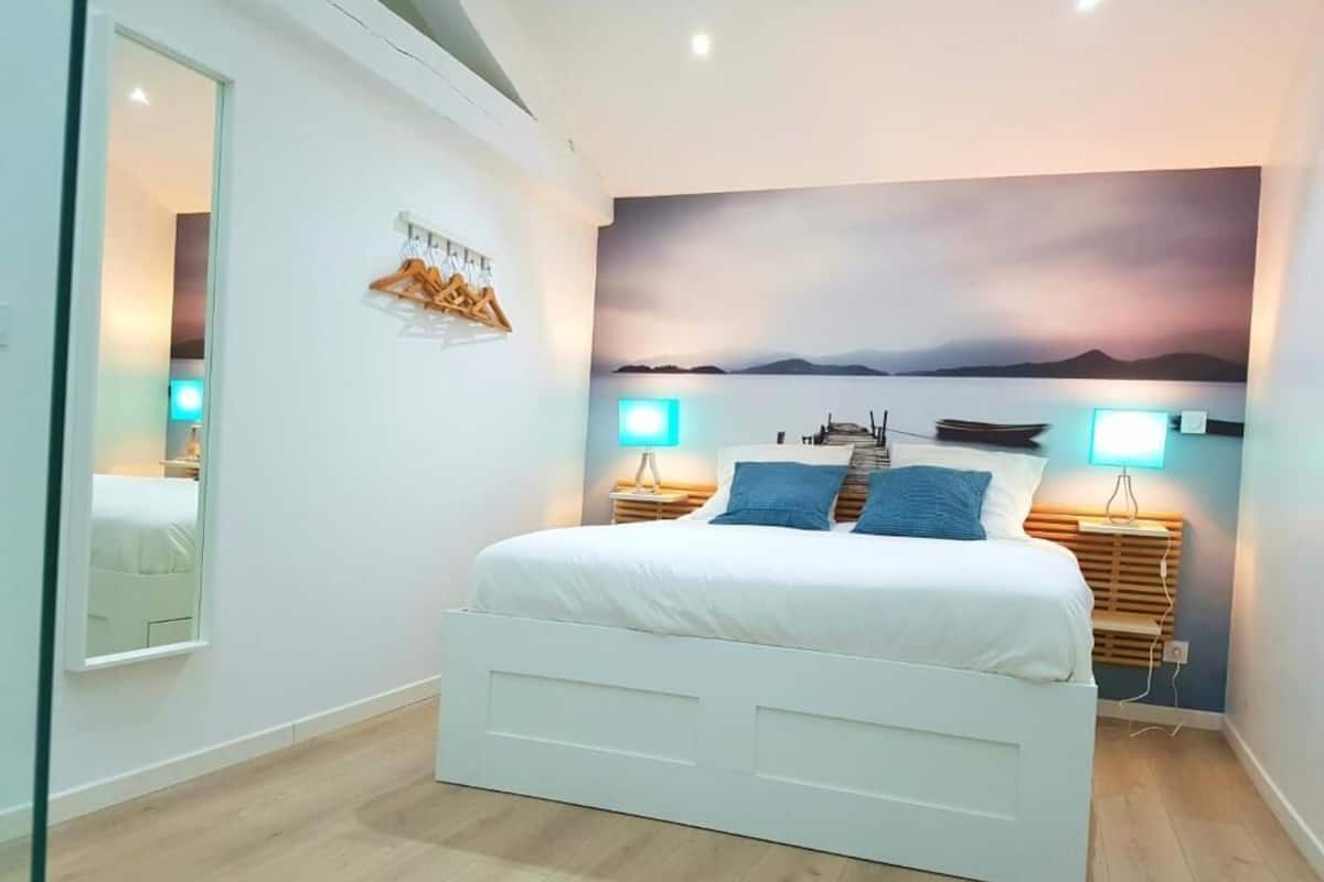 A bedroom with a white bed, wooden headboard, and a mural of a sunset over water. There are two bedside tables with lamps.