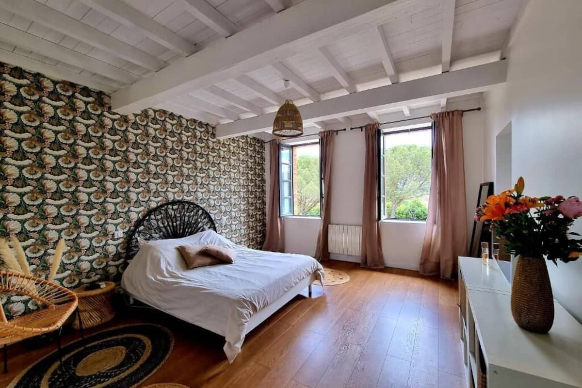 A bedroom with a bed, a wicker chair, a bedside table, a vase with flowers, and a window with curtains.