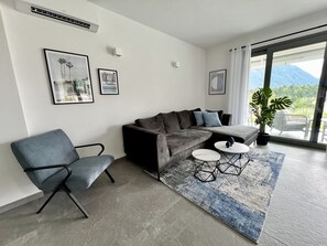 Ein modernes Wohnzimmer mit einem grauen Ecksofa, einem blauen Sessel und einem kleinen Couchtisch. An der Wand hängen zwei gerahmte Bilder und am Fenster steht ein Blumentopf.