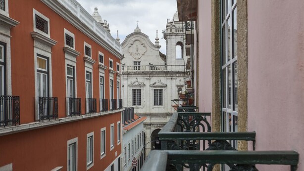 A narrow street with buildings on either side, featuring balconies with wrought iron railings and a prominent white building with a dome and cross. - C2 Authentic Stay | XVIII Century Charm | DT (Lisboa ciudad)