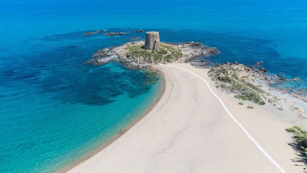 Eine Luftaufnahme einer Küstenregion mit einem Sandstrand, einer kleinen Felseninsel mit einem Turm und klarem, blauem Wasser.