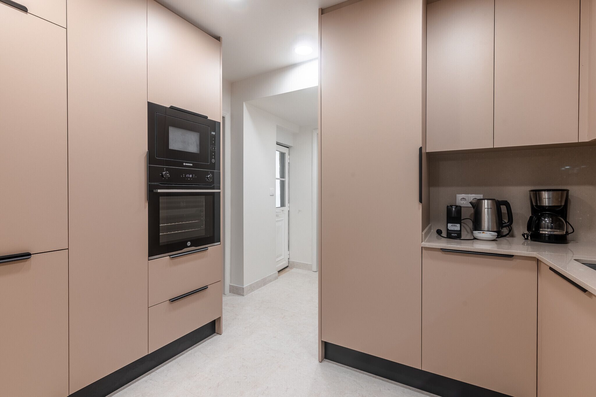 Een moderne keuken met inbouwapparatuur, een aanrecht met een koffiezetapparaat, en een gang die naar een andere kamer leidt.