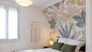 A neatly made bed with a checkered blanket, a wall with a leaf pattern, and a hanging dreamcatcher. - Holiday Home 'L'escale Kénanaise' with Private Garden and Wi-Fi (Saint-Quay-Perros)